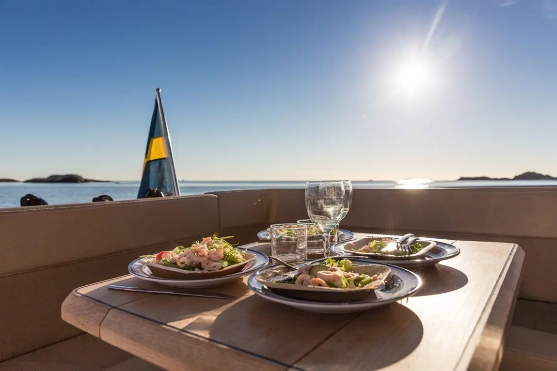 Plates of shrimp salad and glasses on a boat deck, with a view of the sea and sunlit horizon. Swedish flag visible.